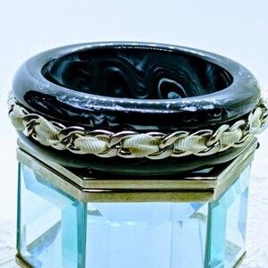 Könnte beinhalten: Ein Stapel schwarzer und weiß marmorierter Harzarmreifen mit einer goldfarbenen Kette und cremefarbenen Stoffdetails. Die Armreifen sind auf einem transparenten Glas-Display gestapelt, mit einem cremefarbenen Band an der Seite.