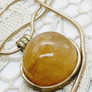 May include: A gold-toned necklace featuring a large, round pendant with a translucent, amber-colored stone. The pendant is set in a gold frame and hangs from a delicate, snake chain. The necklace is displayed on a white, lace-like surface.