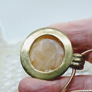 May include: A close-up of a pendant necklace. The pendant has a circular, gold-colored frame with a light-colored, textured stone center. The necklace chain is a thin, copper-colored, snake chain.