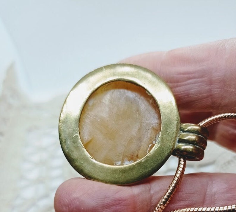 May include: A close-up of a pendant necklace. The pendant has a circular, gold-colored frame with a light-colored, textured stone center. The necklace chain is a thin, copper-colored, snake chain.