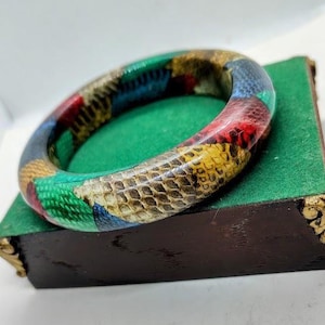 Op de afbeelding: Een kleurrijke armband met een slangenleerpatroon. De armband heeft een mix van kleuren, waaronder rood, geel, blauw en groen. De armband ligt op een groene fluwelen ondergrond.