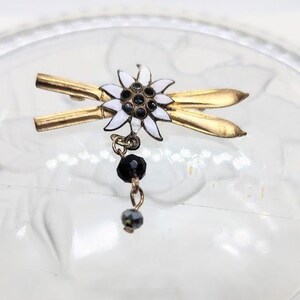 May include: A gold-toned metal brooch featuring a white and black flower design with a dangling black bead accent. The brooch has a pin closure and is set against a blurred background.