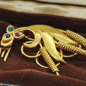 Peut inclure: Broche dorée avec un motif floral, ornée de trois pierres précieuses vertes, rouges et bleues. La broche présente un design texturé et superposé avec une fermeture à épingle, présentée dans un écrin en velours marron.