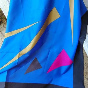 Può includere: Una sciarpa blu con forme geometriche dorate, nere e rosa. La parola "CLANDESTINE" è stampata in bianco. La sciarpa è drappeggiata su una panca bianca.