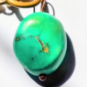 Pode incluir: Um pingente de pedra polida, arredondada e de cor verde brilhante com veios naturais de matriz castanha. A pedra está suspensa por um anel de metal dourado e um elo de corrente a condizer, criando um componente de joalharia orgânico.