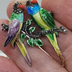 May include: A vibrant parrot brooch depicting two birds on a branch. The birds are painted in green, blue, yellow, and purple with red beaks. The branch and leaves have silver detailing.