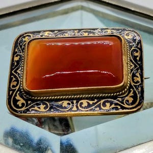 May include: Antique rectangular brooch featuring a polished, reddish-orange stone. The stone is set in a gold-toned frame with a black and gold filigree border. The brooch has a pin clasp on the reverse.