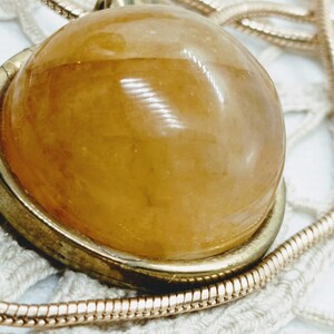 May include: A close-up of a pendant necklace featuring a large, round, amber-colored stone set in a gold-toned metal frame. The pendant hangs from a delicate, gold-colored chain, resting on a white lace surface.