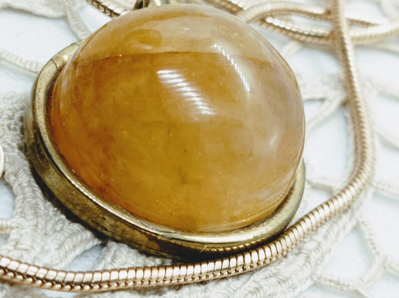 May include: A close-up of a pendant necklace featuring a large, round, amber-colored stone set in a gold-toned metal frame. The pendant hangs from a delicate, gold-colored chain, resting on a white lace surface.