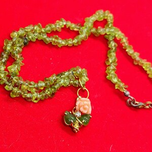 Könnte beinhalten: Eine Halskette mit einer Kette aus kleinen, hellgrünen Peridotsteinen. Ein goldfarbener Anhänger mit einer korallenfarbenen Rose, grünen Blättern und einem kleinen, klaren Edelstein hängt an der Halskette. Der Hintergrund ist einfarbig rot.