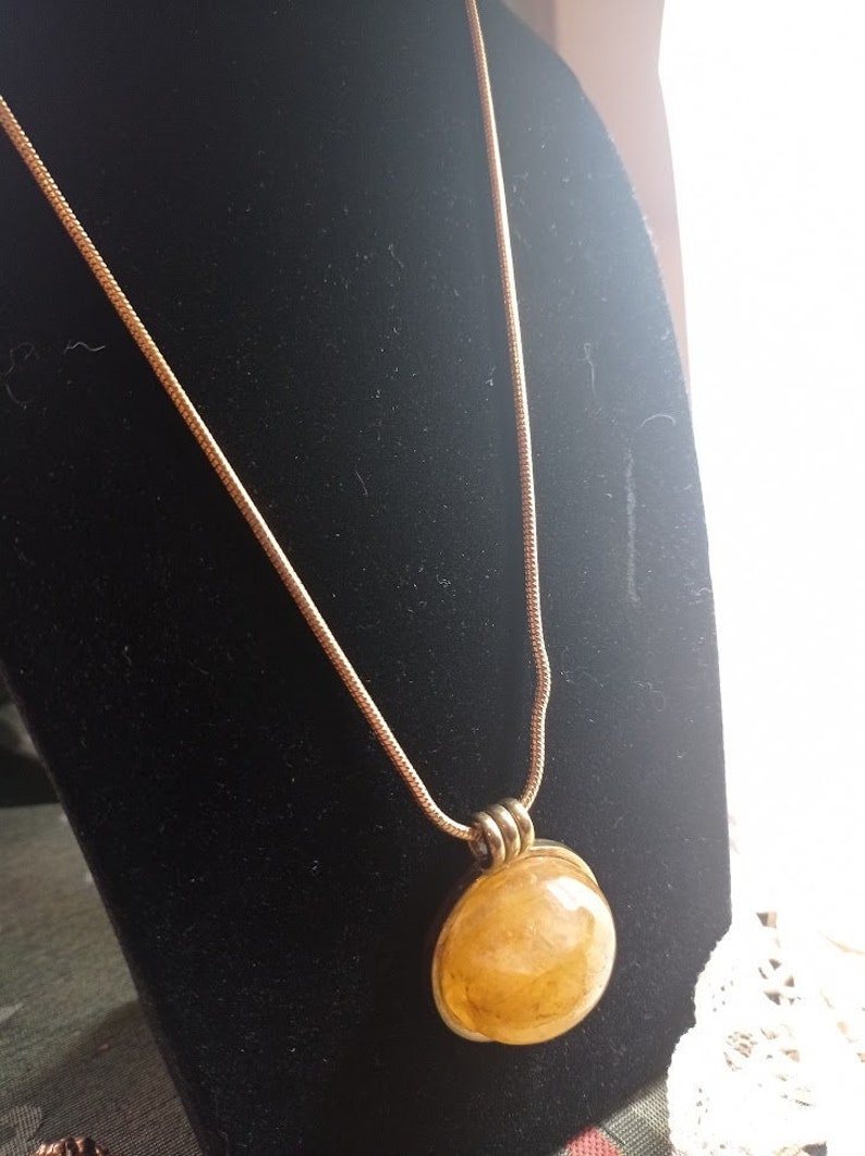 May include: A gold-toned necklace with a round, amber-colored pendant. The pendant is encased in a silver-colored metal setting and hangs from a delicate chain. The necklace is displayed on a black velvet stand.