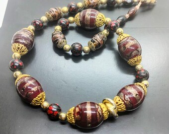 May include: A necklace featuring large, oval-shaped dark red glass beads with gold-toned glitter swirls, interspersed with smaller black beads with red speckles and metallic gold-toned spacer beads, all accented by ornate gold-toned metal bead caps.