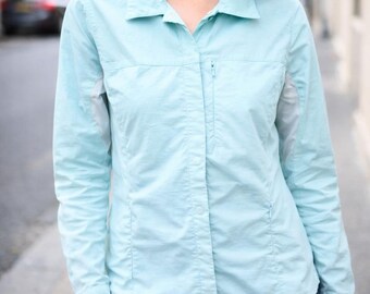 May include: A light blue long-sleeve shirt with a collared neckline and a button-down front. The shirt has a small zipper pocket on the chest and a tailored fit. The sleeves are long and cuffed. The shirt is paired with blue jeans.