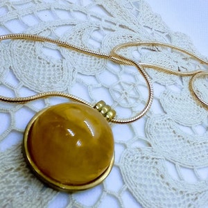 May include: A gold-toned necklace with a large, round pendant. The pendant has a golden-yellow, polished stone set in a gold-colored frame. The chain is a thin, snake-style design. The necklace rests on a white, lace doily.
