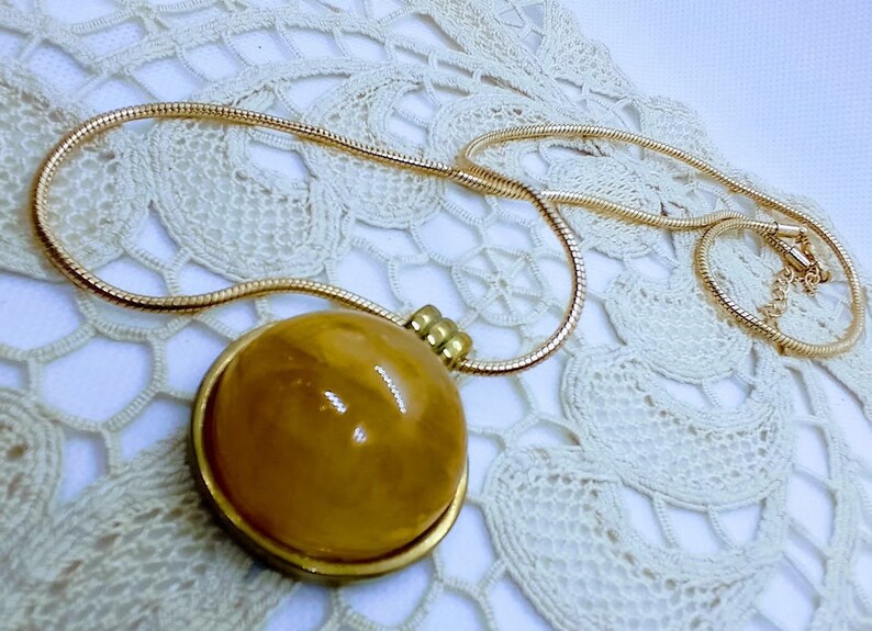 May include: A gold-toned necklace with a large, round pendant. The pendant has a golden-yellow, polished stone set in a gold-colored frame. The chain is a thin, snake-style design. The necklace rests on a white, lace doily.