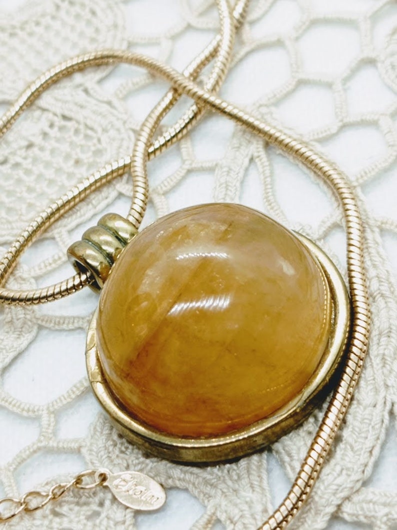 May include: A gold-toned necklace with a large, round, amber-colored pendant. The pendant is set in a gold frame and hangs from a delicate gold chain. The necklace is displayed on a white, lace-like surface.