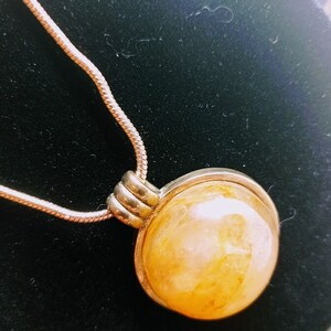 May include: A silver-toned necklace featuring a large, round, amber-colored pendant. The pendant is encased in a silver metal frame and hangs from a delicate chain. The pendant has a marbled appearance with variations in color.