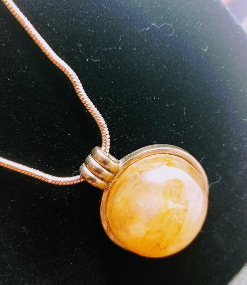 May include: A silver-toned necklace featuring a large, round, amber-colored pendant. The pendant is encased in a silver metal frame and hangs from a delicate chain. The pendant has a marbled appearance with variations in color.