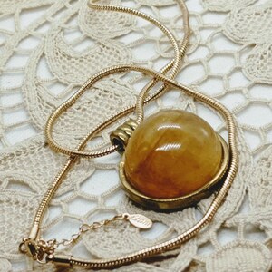 May include: A gold-toned necklace with a large, round, amber-colored pendant. The pendant is set in a gold-colored bezel and hangs from a delicate, snake chain. The necklace is displayed on a cream-colored, lace-like surface.