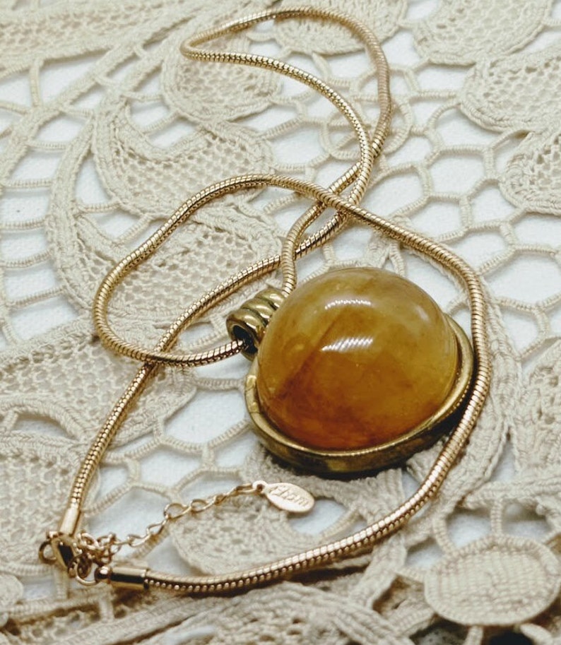 May include: A gold-toned necklace with a large, round, amber-colored pendant. The pendant is set in a gold-colored bezel and hangs from a delicate, snake chain. The necklace is displayed on a cream-colored, lace-like surface.