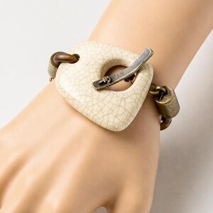 Op de afbeelding: Een crèmekleurige vierkante armband met een craquelé-afwerking en een zilverkleurige sluiting. De armband heeft bronskleurige schakels en een cilindervormige bedel. De armband wordt om de pols gedragen.