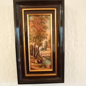 May include: A framed landscape painting depicting a tranquil scene with trees, a small house, and water. The artwork is set within a dark brown frame with a gold inner border, offering a traditional look.