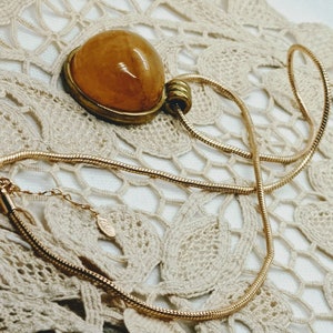 May include: A gold-toned necklace with a pendant featuring a large, oval, amber-colored stone. The pendant is set in a gold-colored metal frame. The necklace chain is a thin, snake chain style, and the background is a cream-colored lace.