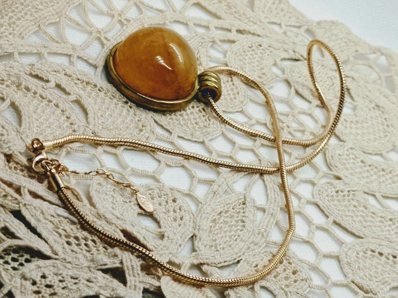 May include: A gold-toned necklace with a pendant featuring a large, oval, amber-colored stone. The pendant is set in a gold-colored metal frame. The necklace chain is a thin, snake chain style, and the background is a cream-colored lace.