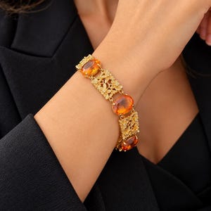 Peut inclure: Un bracelet doré composé d'une alternance de pierres précieuses ovales orange et de maillons rectangulaires ornés de motifs floraux en filigrane. Le bracelet est constitué d'une série de segments métalliques reliés entre eux.
