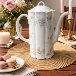 Op de afbeelding: Een witte porseleinen koffiepot met een deksel, versierd met een delicaat bloemmotief. De pot staat op een geweven placemat op een houten tafel, samen met kaarsen, bloemen en macarons. De pot is ongeveer 25 cm hoog.