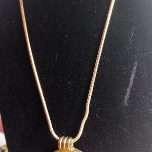 May include: A gold-toned necklace with a round, light-yellow pendant. The pendant is encased in a silver-colored metal frame. The chain is a thin, snake-style design. The necklace is displayed against a black background.