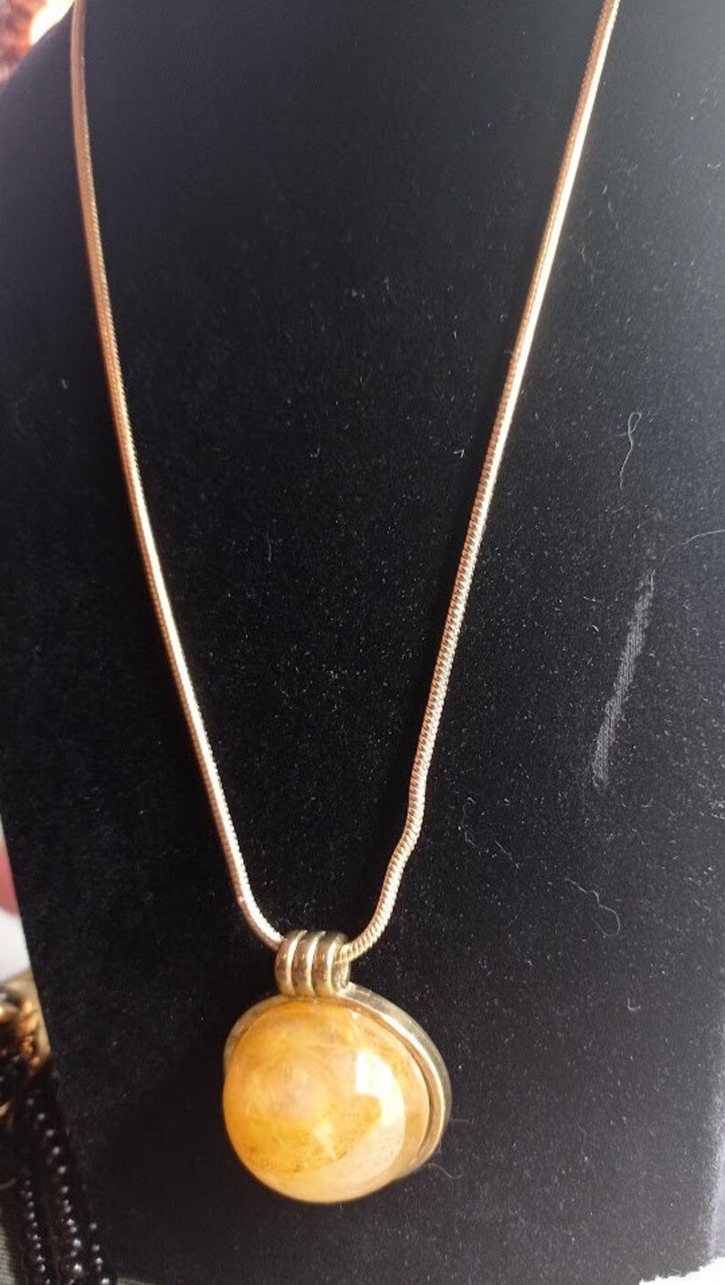 May include: A gold-toned necklace with a round, light-yellow pendant. The pendant is encased in a silver-colored metal frame. The chain is a thin, snake-style design. The necklace is displayed against a black background.