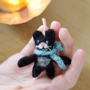 Könnte beinhalten: Handgefertigter schwarzer Katzenanhänger mit hellblauem Schal. Die Katze hat ein weißes Gesicht und eine weiße Brust und wird an einer weißen Schnur aufgehängt. Der Anhänger wird in der Hand einer Person gehalten, mit einer brennenden Kerze im Hintergrund.