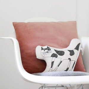 May include: A white chair with a pink pillow and a black and white cat pillow. The chair is in a white room with a white lamp.