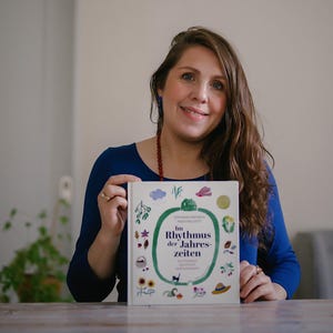 May include: A woman holding a book titled "Im Rhythmus der Jahreszeiten." The book cover is white with colorful illustrations of nature elements. The woman is wearing a blue long-sleeved shirt and a red beaded necklace.