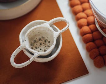 May include: White ceramic tea strainer with two handles. The strainer is nested inside a white ceramic bowl.