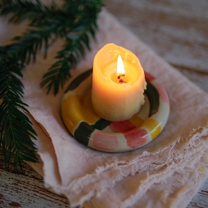 Può includere: Una candela accesa su un portacandele in ceramica colorata con un motivo a strisce. La candela e il portacandele poggiano su un panno rosa chiaro sfilacciato, con un rametto di verde sullo sfondo. La fiamma della candela brilla.
