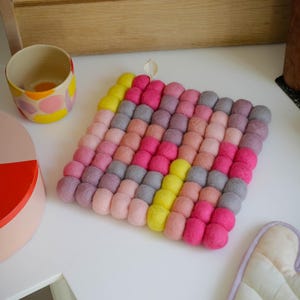 Könnte beinhalten: Ein quadratischer Filz-Topfuntersetzer mit Aufhängeschlaufe. Der Untersetzer besteht aus kleinen, runden Filzkugeln in Rosa-, Gelb- und Grautönen. Ein passender Ofenhandschuh ist ebenfalls zu sehen.