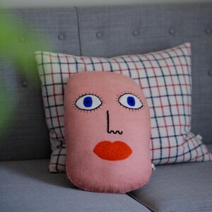 Könnte beinhalten: Ein rosa Zierkissen mit einem skurrilen Gesichtsdesign mit blauen Augen, einer schwarzen Nase und einem orangefarbenen Mund. Das Kissen liegt auf einem grauen Sofa mit einem karierten Kissen im Hintergrund.