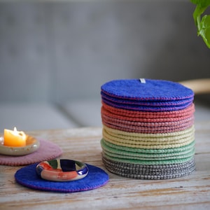 Könnte beinhalten: Ein Stapel bunter Filzuntersetzer in Blau-, Rosa-, Gelb-, Grün- und Grautönen. Ein blauer Untersetzer mit einem dekorativen Keramikring und ein violetter Untersetzer mit einer brennenden Kerze sind ebenfalls zu sehen. Die Untersetzer stehen auf einem Holztisch.