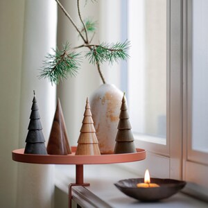 Op de afbeelding: Een set van vijf houten kerstboomversieringen in verschillende tinten bruin en grijs, gerangschikt op een klein, rond, terracottakleurig dienblad. Het dienblad is met een klem aan een vensterbank bevestigd. Een kleine, brandende kaars staat in een bruine schaal op de vensterbank.