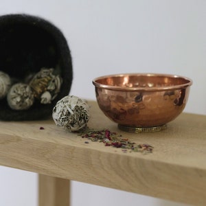 May include: A copper bowl with a hammered texture sits on a wooden table with dried herbs and white sage bundles.