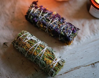 Può includere: Due mazzi di erbe secche legate con spago, uno con erbe viola e verdi, l'altro con erbe gialle e verdi. Sullo sfondo un cristallo trasparente con una candela accesa all'interno e un barattolo marrone con una candela all'interno.