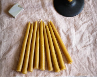 May include: A set of eight yellow beeswax taper candles on a light pink linen fabric. The candles are arranged in a fan shape. A small white box and a black candle holder are also visible in the image.