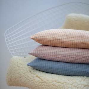 May include: Three checkered throw pillows in shades of pink and blue are stacked on a white sheepskin throw resting on a white wire chair.