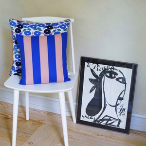 Könnte beinhalten: Ein weißer Stuhl mit einem rosa-blauen gestreiften Kissen auf der Sitzfläche. Ein schwarz-weiß gerahmtes Bild eines Gesichts lehnt an dem Stuhl.