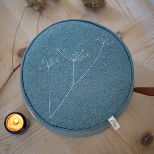 Könnte beinhalten: Ein rundes Meditationskissen in Teal mit einem weißen, gestickten Blumendesign. Das Kissen hat einen braunen Ledergriff und ein weißes Etikett mit der Aufschrift "Otter."