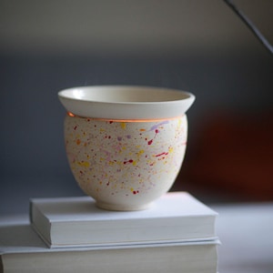 Könnte beinhalten: Eine weiße Keramikschüssel mit einem gesprenkelten Muster aus gelber, rosa und roter Farbe. Die Schüssel steht auf einem Stapel Bücher.