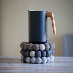 Könnte beinhalten: Ein schwarzer Wasserkocher mit Holzgriff steht auf einem Stapel grauer und brauner Filzkugeln. Auf dem Wasserkocher ist der Schriftzug "Smeg" aufgedruckt.