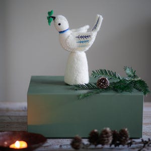 Op de afbeelding: Een witte vilten duif figuur met blauwe geborduurde details en een groen blad in zijn bek. De duif staat op een witte basis, bovenop een groene doos. Decoratieve dennenappels en groen zijn in de buurt geplaatst.